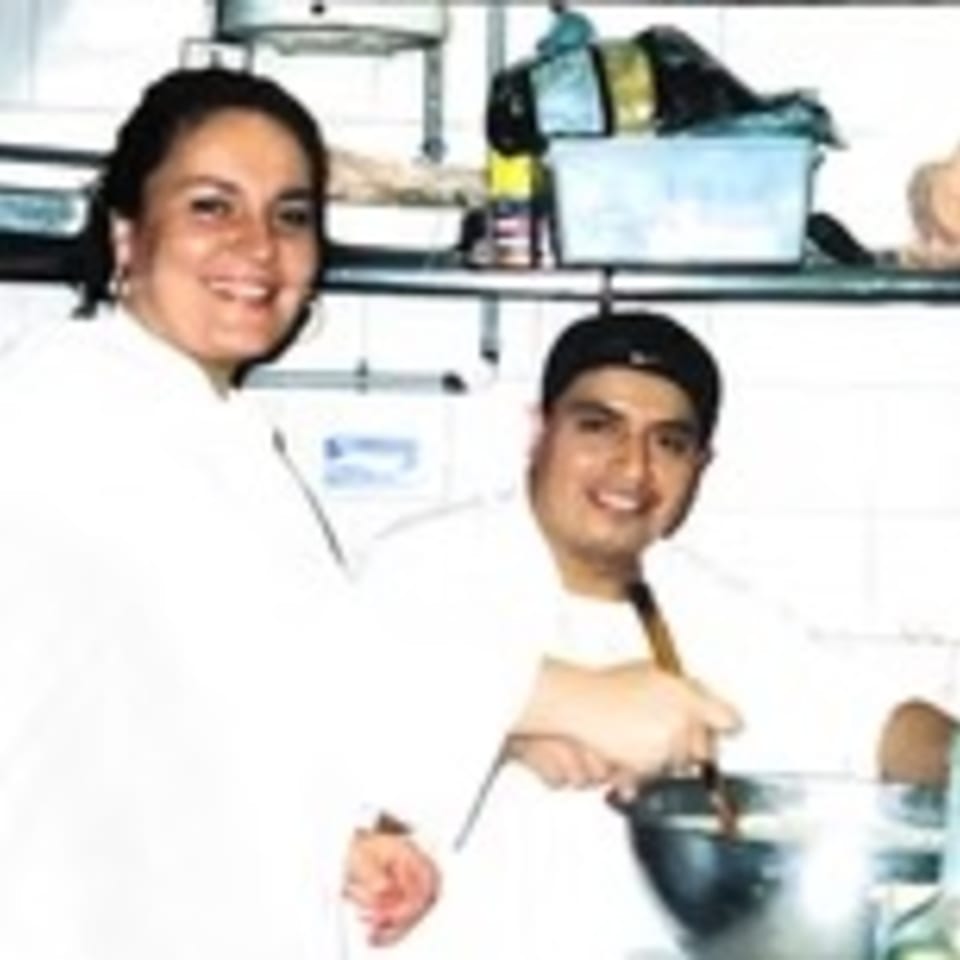 Two smiling chefs in white uniforms, one stirring a pot, in a busy kitchen.