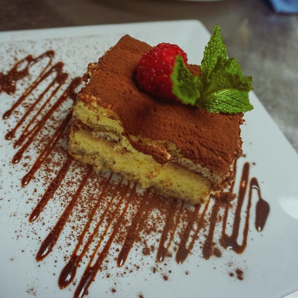 A slice of tiramisu with cocoa powder, a raspberry, and mint on a white plate.