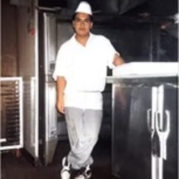 A young chef in a white uniform and hat stands in a commercial kitchen.