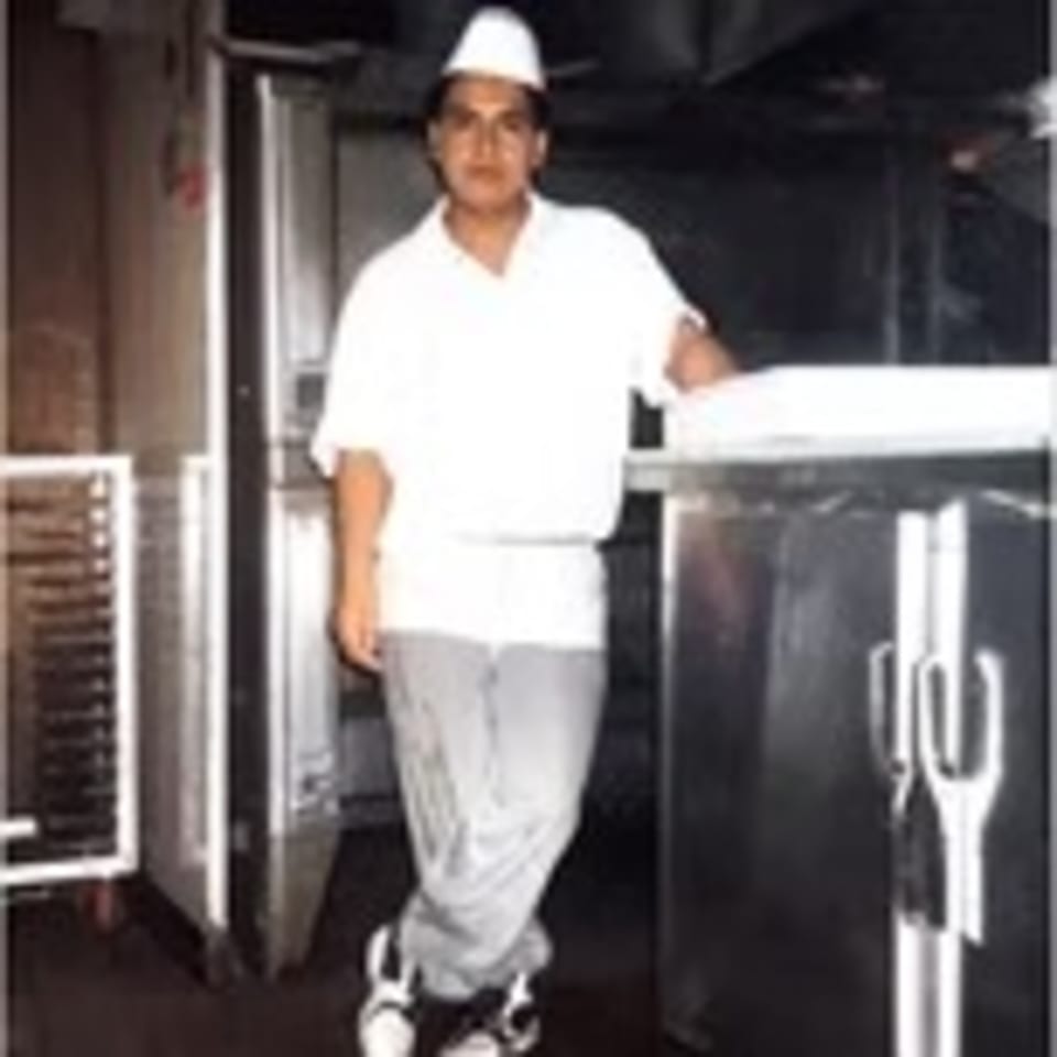 A young chef in a white uniform and hat stands in a commercial kitchen.